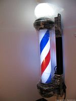 Front view of the illuminated barber pole mounted on a wall, wrapped with protective plastic film. Shows the red, white, and blue spiral pattern brightly lit inside a clear acrylic cylinder, topped with a glowing ball light.