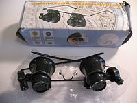 Box and glasses with dual magnifying lenses and LED lights seen resting in plastic, showcasing the packaging that references glasses type watch repair magnifier with LED light 20x magnification.