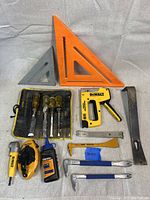 Overview of hand tool lot showing two carpenter's squares, set of chisels in tool roll, DeWalt staple gun, pry bars, chalk line, oil bottle, rotary tool, and scrapers