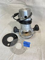 Front and top view of Craftsman router with attached black cord and two round accessories: clear plastic base plate and black mounting plate.