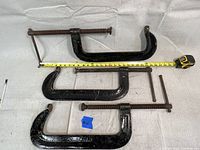 Three large black metal C-clamps laid flat, each showing approximate size with tape measure reaching nearly 27 inches in total length including screws.