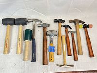 Full view of 12 hammers arranged on white cloth showing variety of hammer types and handle materials.