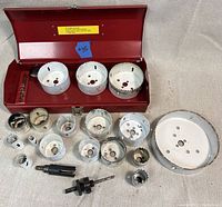 Wide view of lot showing many white hole saws of various sizes and red Milwaukee metal case with three large hole saws inside and multiple hole saw components and arbors in front.