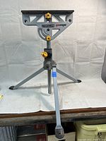 Front view showing full tripod legs, height adjustment, and flat clamp surface with brand and model markings.