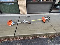 Full side view of the STIHL FS 90 gas-powered brush cutter and trimmer showing the engine, handlebar with throttle control, long shaft, and cutting head with orange shield.