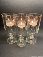 View of three glass candle holders each lit with a tea light candle, showing the delicately blown shapes inside tubular vessels.