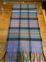 Full view of the scarf rolled at one end showing the check pattern in grey/blue, muted green, tangerine, and black with twisted fringe ends.