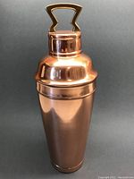 Full view of copper cocktail shaker with brass handle on grey background, showing overall polished copper finish and lid fit.