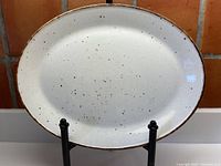 Front view of an oval ceramic platter with cream glaze and cinnamon speckles, supported on display stand.