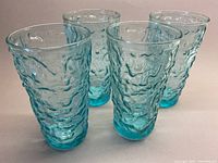 Four turquoise glass tumblers with a highly textured crinkle pattern on their exterior, shown from a front angle.