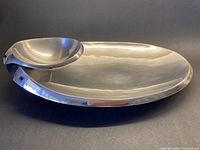 Side view of the oval concave metal chip and dip bowl, showing the attached smaller dip bowl held by a riveted black bar.