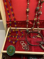 Photo showing red velvet-lined box with assorted charms, bracelets, copper cuff, and necklaces including cloisonné bead necklace and seed bead bracelet.