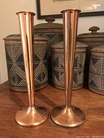 Photo showing pair of vintage copper candlesticks in front of decorative jars on a wooden surface, highlighting shape and size.