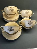 Full view of the set showing 8 double-handled bowls stacked with 8 matching saucers underneath; bowls are cream colored with gold floral basket motif.
