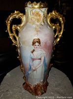 Front view of antique porcelain vase showcasing a hand-painted lady in white dress with blue drape and a red waistband, gilt handle detail.