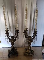 Pair of vintage heavy brass candelabras with tall ornate taper candles, crystal accents hanging from the arms, placed on marble surface against cream wall.