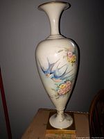 Tall slender hand painted porcelain vase with bluebird and flower motifs, shown against dark background with cardboard filler at base.