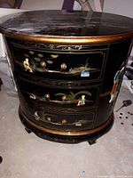 Front view of the round lacquered accent table with three drawers, showing the detailed hand-painted Asian motifs with figures and nature scenes.