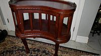 Full view of the polished oval wooden display table showing glass panel doors, curved legs, and decorative apron.