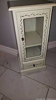 Front view of small cream accent storage cabinet with glass door and bottom drawer displaying leaf pattern decoration.