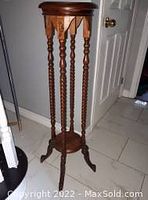 Photo shows tall antique wooden plant stand with spindle legs, circular top and bottom shelves on tiled floor next to closed white door.
