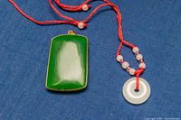 Photo showing both jade pendants on red cords clearly, rectangular green jade with gold-tone frame and white jade circular coin shape with beads.