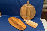 Three wooden cutting and cheese boards on a blue cloth background: one round with handle and signature, one oblong, and one small rectangular with handle and string.