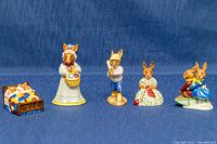 Five Royal Doulton Bunnykins porcelain figurines including a bunny in bed, Mrs. Bunnykins with basket, baseball player, female bunny in floral dress, and two dancing bunnies, arranged on blue background.
