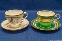 Two Aynsley bone china teacup and saucer sets, one white with skier and text, the other green with gold pattern