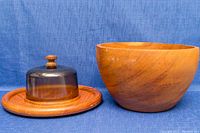 Cheese board with glass dome cover and teak salad bowl shown side by side on blue fabric background.