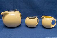 Photo shows the front view of the three-piece tea set with lidded teapot, lidded sugar bowl, and creamer on blue fabric background.