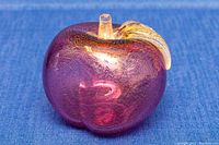 Front and side view of purple Murano apple paperweight with golden fleck leaf and clear stem on blue fabric.