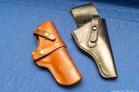 Photo showing both black and brown leather gun holsters side by side on a blue fabric background to display size, color, and general condition.