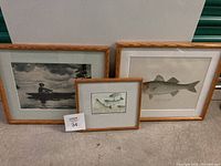 Three framed prints grouped together on floor. Small fish illustration centered between two larger prints: one fish illustration, one fisherman and boat scene.
