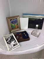 Lot items inside white artist tent showing canvases, framed painting, airbrush kit with accessories, and instruction manual.