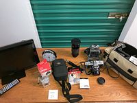 Overview of all items including Canon Powershot S400 digital camera, Minolta SRT 201 camera, Chyron and Canon lenses, Canon filter and cleaning kit, carrying bag, analog clock, and Samsung TV.