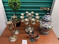 Full view of lot showing two candlesticks, large candelabra, smaller three-candle candelabra, artificial topiary tree, and lidded vase on display surface.