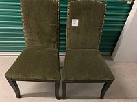 Photo of two dark green velour dining chairs with black wooden legs and nailhead trim, showing fabric texture and overall condition.