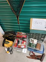 Complete fishing lot arranged on the ground in front of green roll-up door including fishing net, spotlight, tackle boxes, lures, hooks, multi-tool.