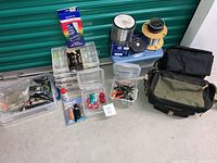 Full view of fishing lot items arranged showing multiple plastic storage containers with fishing lures, fishing reels stacked with spools of line, fishing bags, and small tackle boxes.