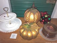Photo showing white two-tier serving dish with ruffled edges and gold handle, cluster of shiny multicolored Christmas ball ornaments, copper lighted pumpkin, smaller gold decorative pumpkin, and stacked orange iridescent party dishes.