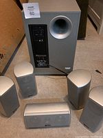 Subwoofer and all five satellite speakers displayed on floor, silver/gray color