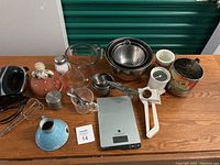 Photo of baking and cooking items arranged on wood surface showing stainless steel mixing bowls, glass measuring cups, hand mixer, kitchen timer, food scale and various baking tools