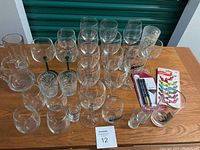 Front shot showing multiple clear stemmed wine glasses, green-stemmed wine glasses, short tumbler glasses, shot glasses, and a colorful beer stein with text. Also includes a pack of black straws and set of cat-themed shot glasses.