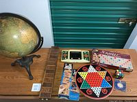 Wide view of the lot showing the freestanding globe, cribbage board, Jenga, Chinese checkers board, Scrabble, Rubik's cube, and electronic football game on a wooden table against a green storage door.