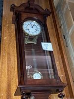 Full view of the mahogany rectangular wall clock showing the arched top, Roman numeral face, brass pendulum, and wood details.