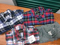 Four men's flannel shirts folded on wooden surface, showing variety of plaid patterns and colors, with auction tag visible.
