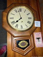 Full view of solid oak Ansonia wall clock with octagonal frame, clock face with Roman numerals, pendulum case, cracked glass, pink instruction sheet and winding key shown beside it.