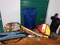 Photo of assortment of sports gear on wooden surface including mesh bag of balls, badminton bag, blue folding sports chair, and racket bags.