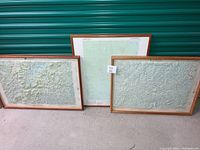 Three framed topographical maps leaning against a green corrugated door on concrete floor. All three maps have wood frames and are approximately 28 x 21 inches each.